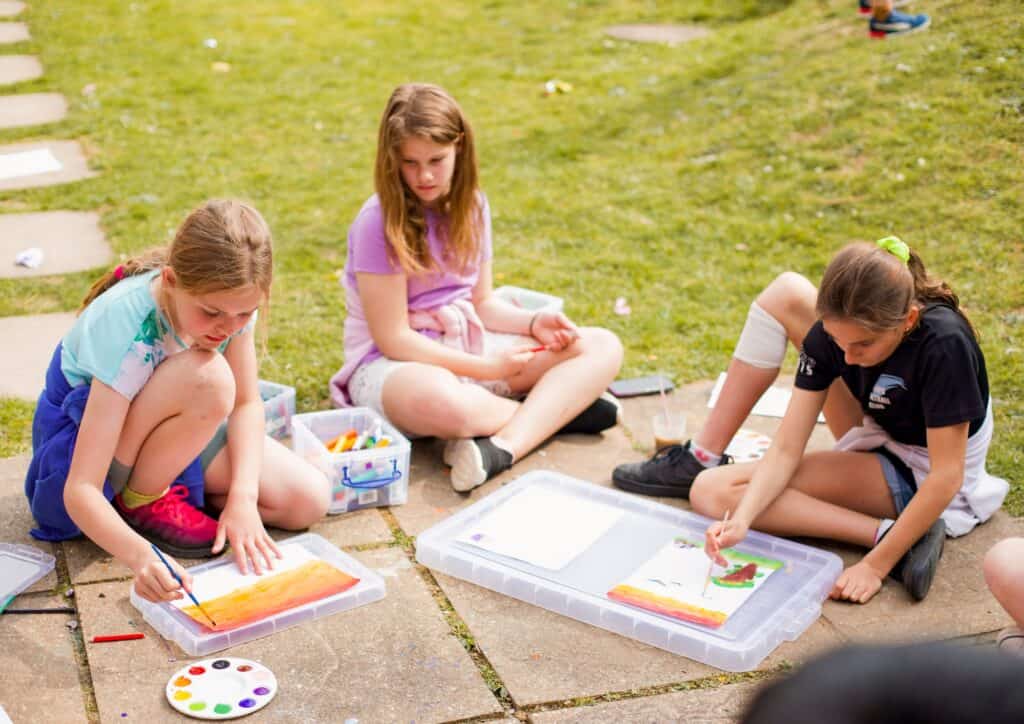 Three smiling girls sitting outdoors under the warm summer sun, enjoying a creative painting activity at Educamps. Surrounded by nature, they’re focused and full of joy as they bring their colourful artwork to life — a perfect sunny day of fun, creativity, and friendship at our summer camp for kids.