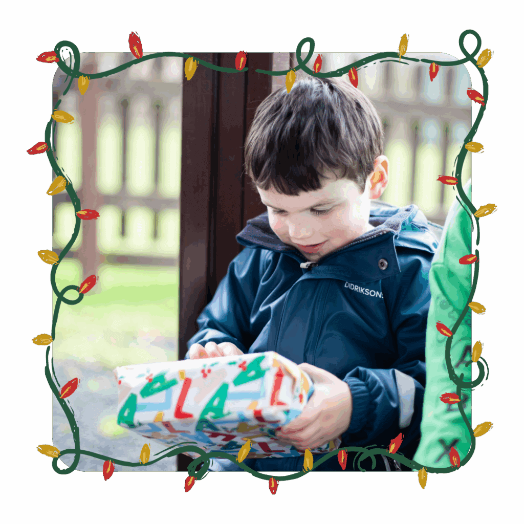 A young boy in a blue raincoat smiling as he opens a Christmas-themed present during an Educamps winter activity.