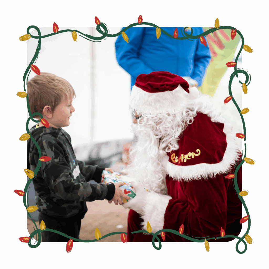 A young child smiling while receiving a wrapped Christmas present from a person dressed as Santa Claus at a festive holiday event.